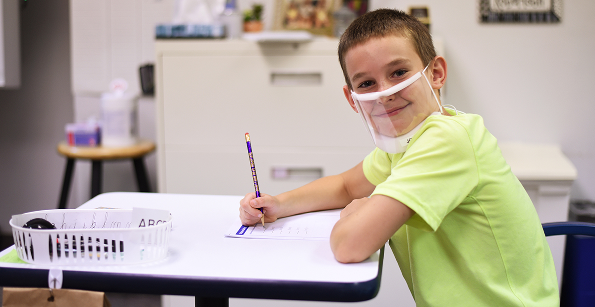 A young boy wearing a face mask is seated at a desk with his materials spread out.