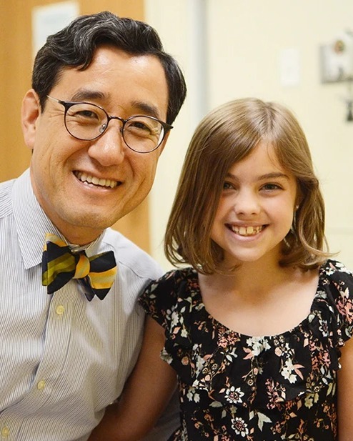 A doctor in a striped bow tie and glasses smiling with a young patient in an exam room.