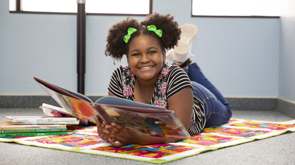 A smiling girl with green hair bows lies on a colorful quilt, reading a large picture book beside a stack of children’s books.