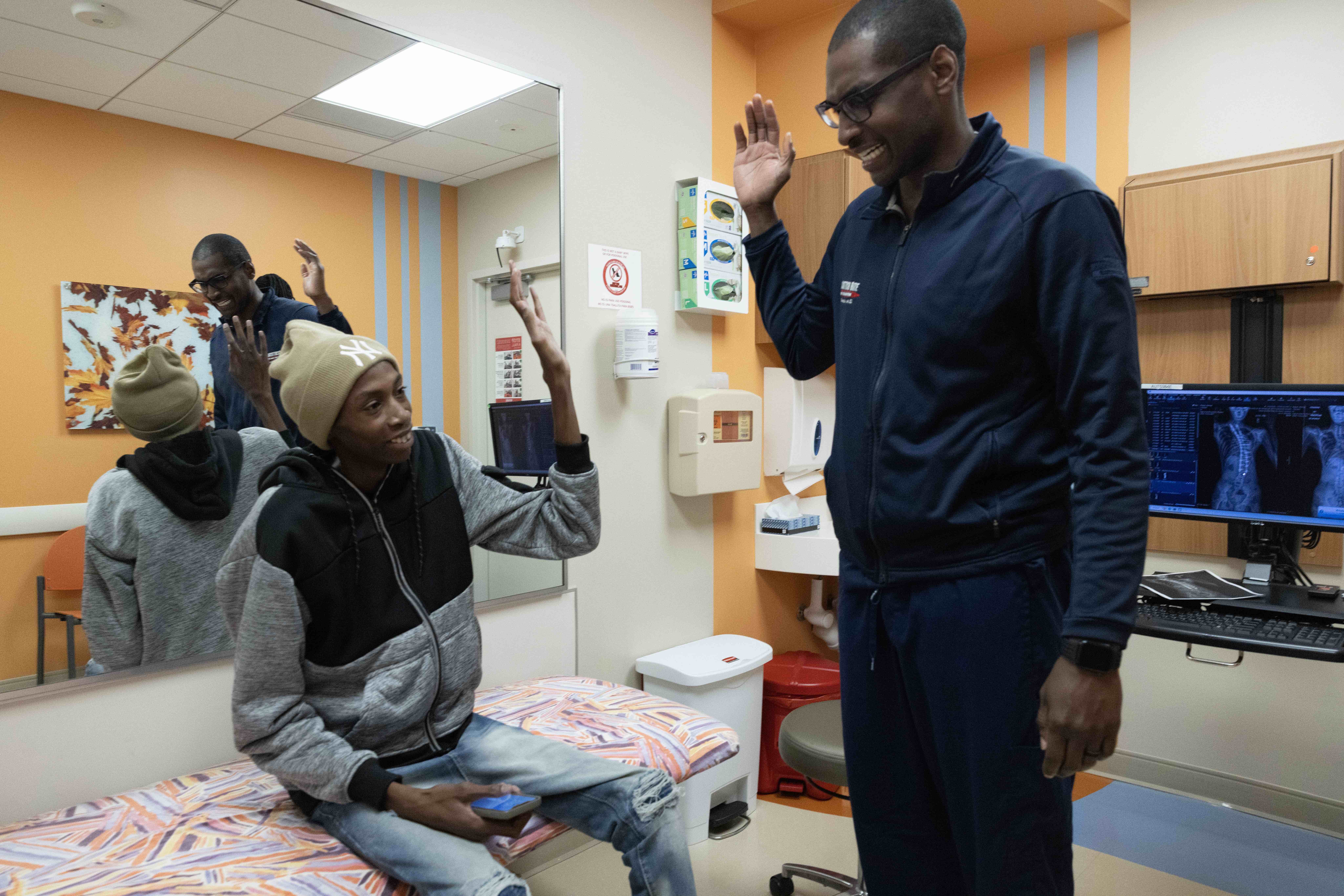A smiling man in glasses and a navy zip-up jacket stands in an orange exam room and raises his hand for a high five with a young person seated on the exam table, who wears a tan beanie and a gray and black hoodie and raises a hand back.