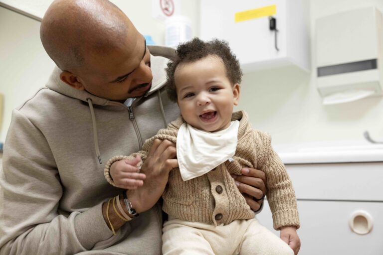 A laughing baby in a tan knit cardigan and white bib sits on a man’s lap in an exam room as the man looks down at the baby.